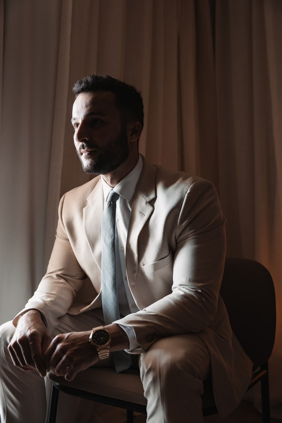 Stylish groom in beige suit and gray tie sitting on a chair by a curtained window looking away.