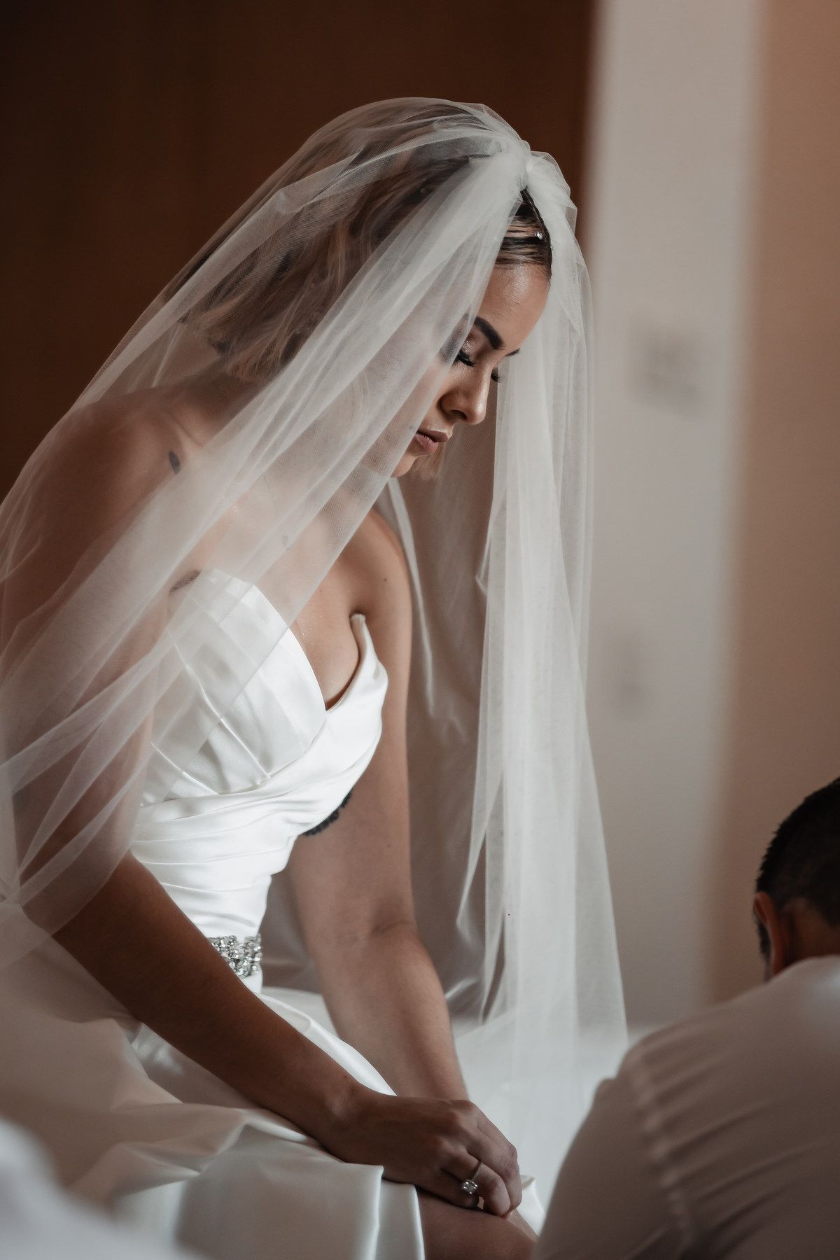 Bride in strapless white gown and flowing veil looking down during wedding preparations.