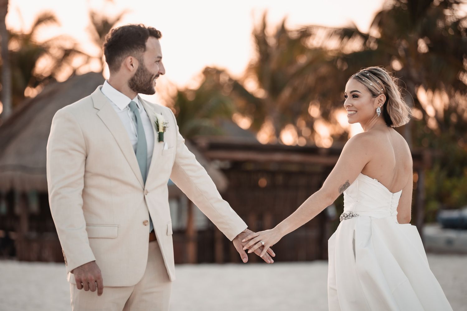 Bride and groom holding hands on sandy beach at sunset during tropical destination wedding celebration.