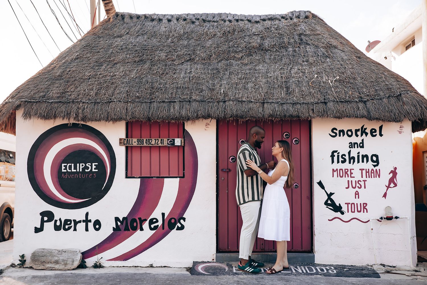 Couple embracing outside a colorful mural wall in Puerto Morelos, Mexico, advertising snorkel and fishing tours.