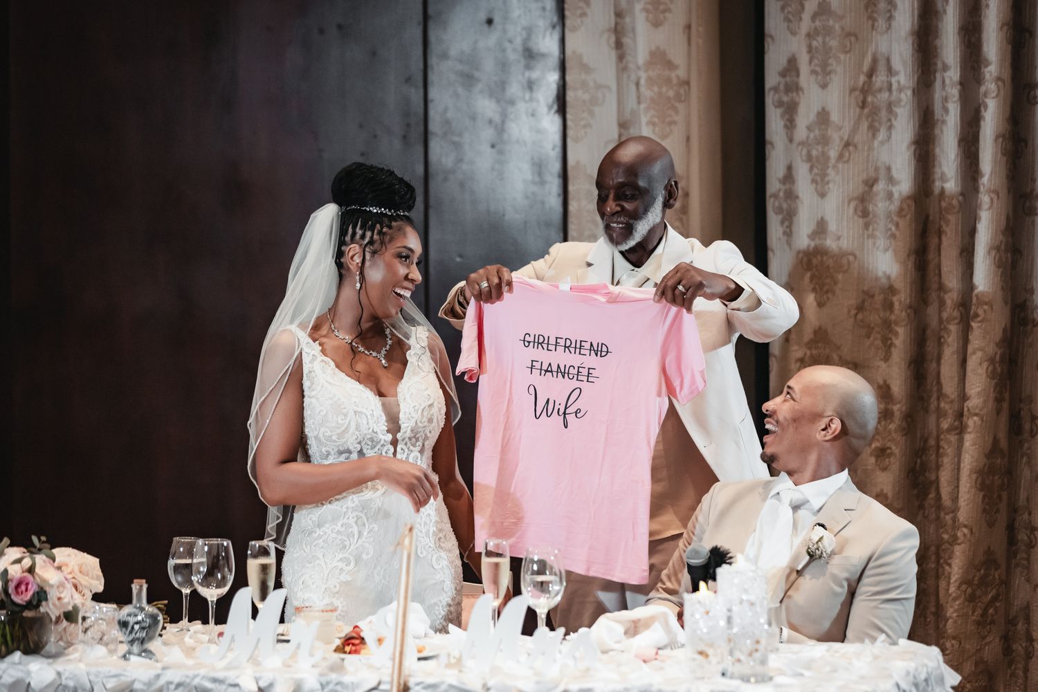 Bride and groom holding a pink shirt reading Girlfriend Fiancee Wife at their wedding reception table.