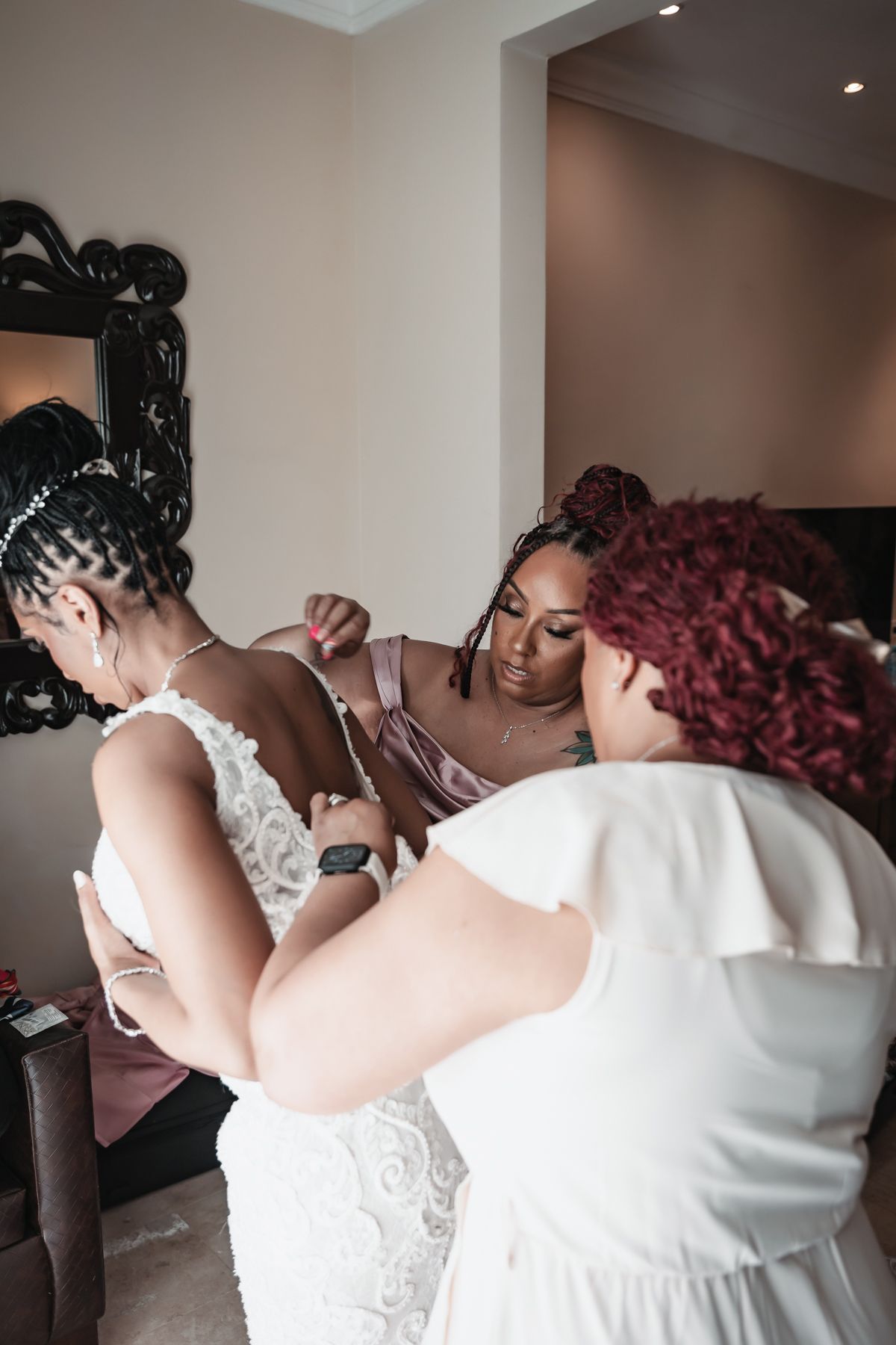 Bridesmaids helping bride into white lace wedding dress in hotel room mirror.
