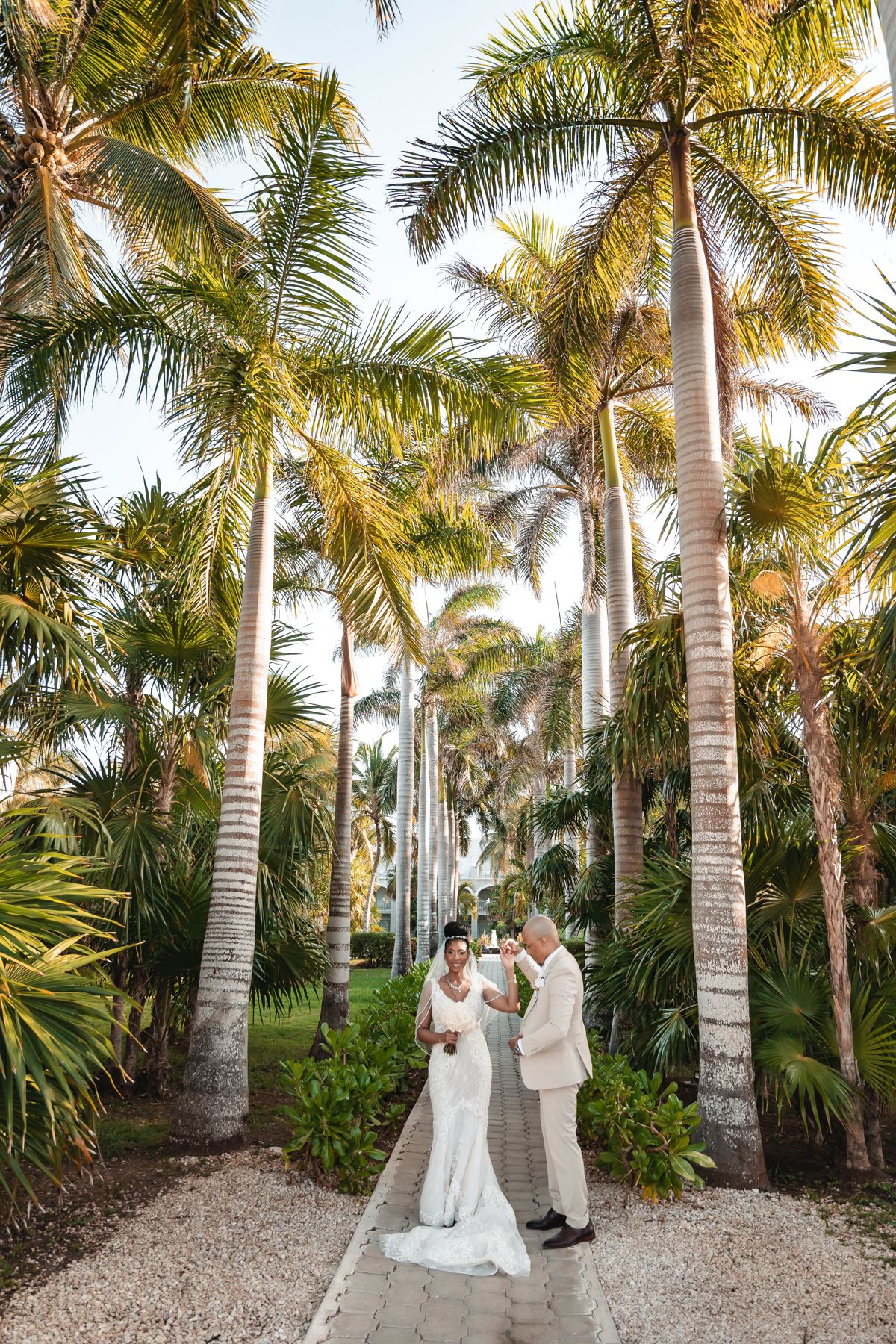 Bride and groom stand on a palm tree-lined path in a tropical garden during their outdoor wedding ceremony.