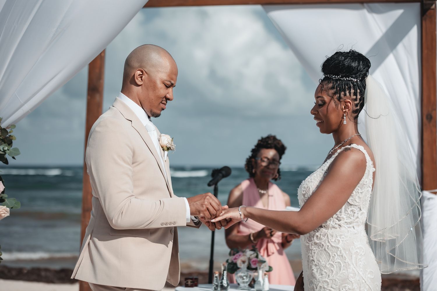 Couple exchanging rings during a beach wedding ceremony with an officiant and ocean backdrop.