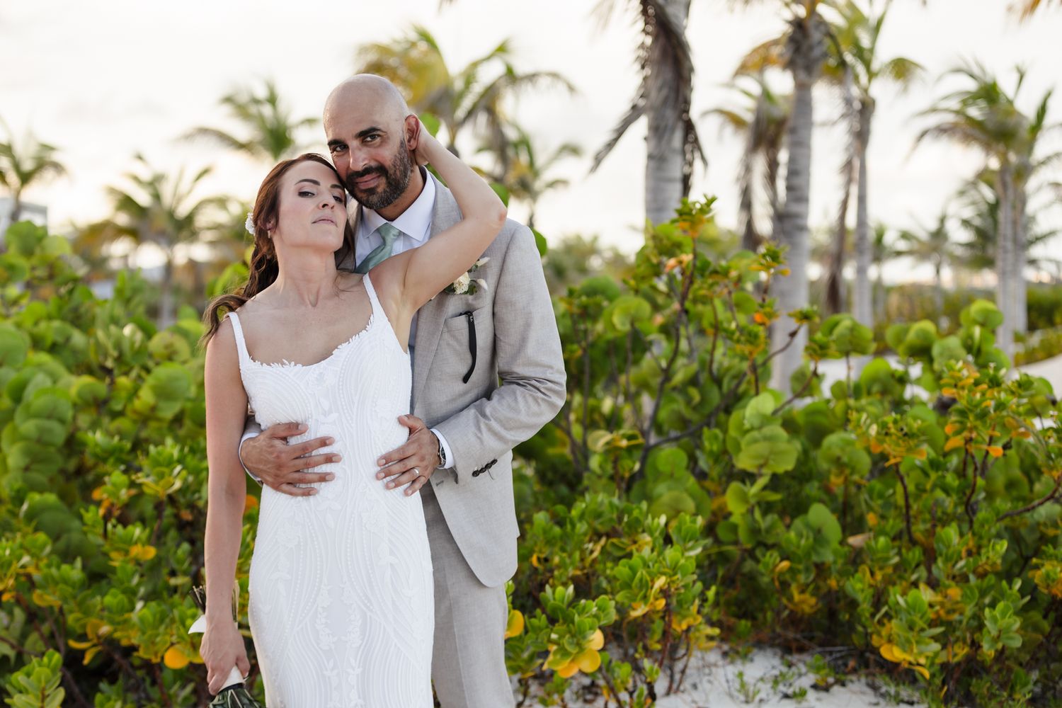 Couple embracing on tropical beach wedding, groom in gray suit holding bride in white dress among palm trees.