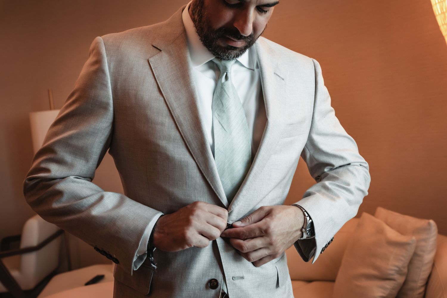 Man in light gray suit adjusting jacket buttons, wearing white dress shirt, light blue tie, and watch.