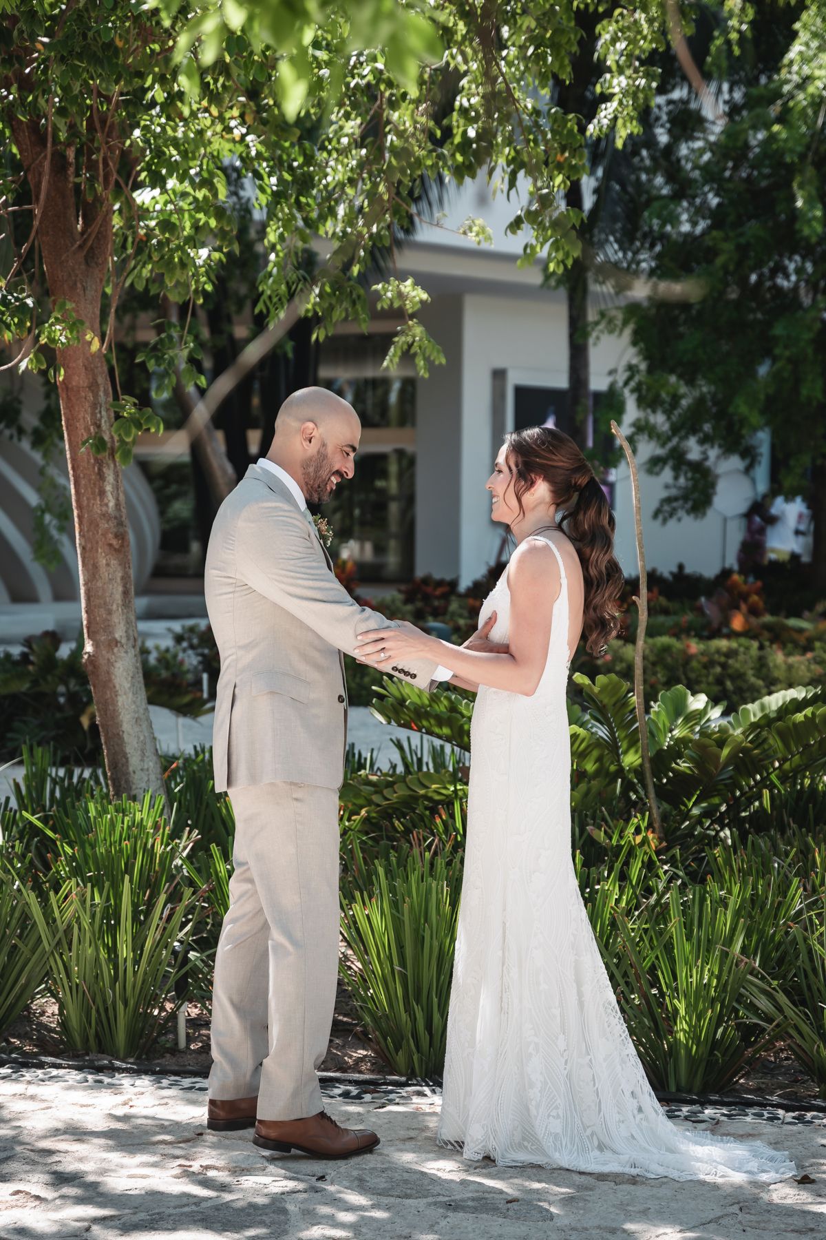 Bride and groom share a tender first look moment outdoors, surrounded by lush greenery near a white building.