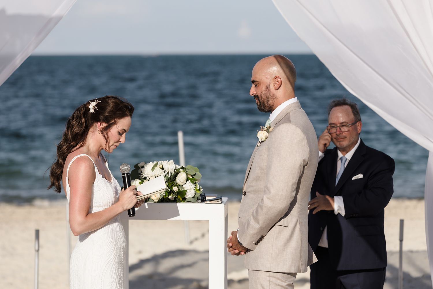 Bride and groom exchange vows at a romantic beach wedding ceremony with an ocean backdrop and white draped arch.