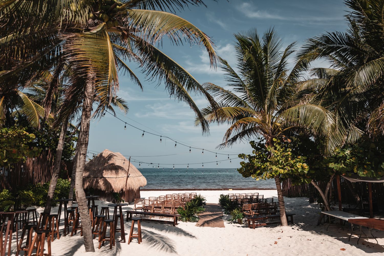 Tropical beach restaurant with palm trees, string lights, and wooden chairs on white sand overlooking a calm turquoise sea.