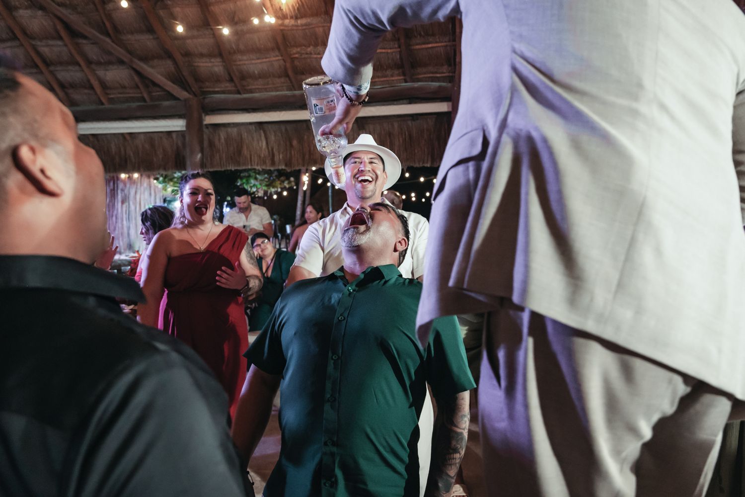 Wedding guests celebrate and dance joyfully inside a rustic venue, with a man in a white hat smiling at the camera.