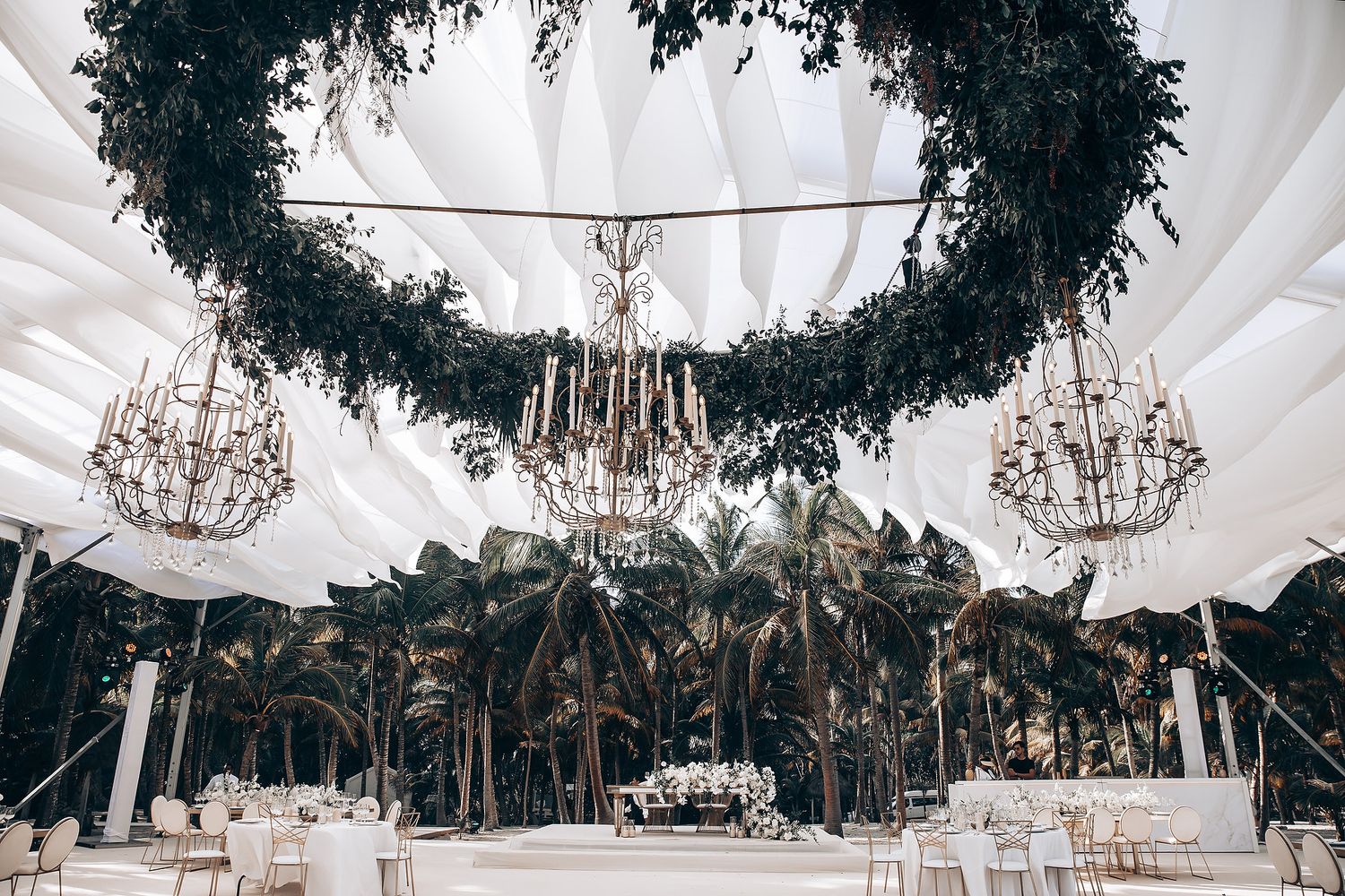 Outdoor wedding venue with draped white canopy, crystal chandeliers and tropical palm trees in background.