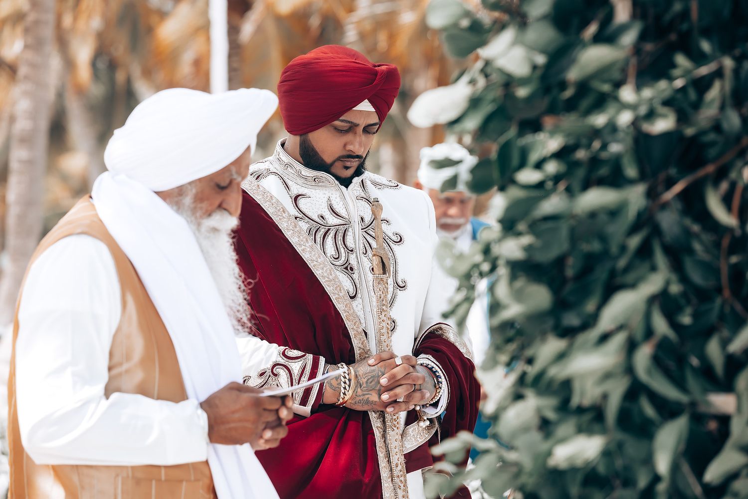 Traditional Sikh wedding ceremony taking place outdoors with guests seated on white cushions under palm trees.
