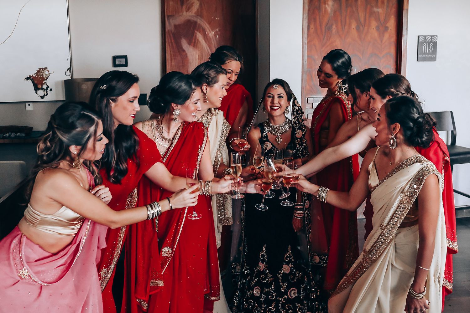 Wedding party members in red and pink saris celebrate together in an indoor setting.