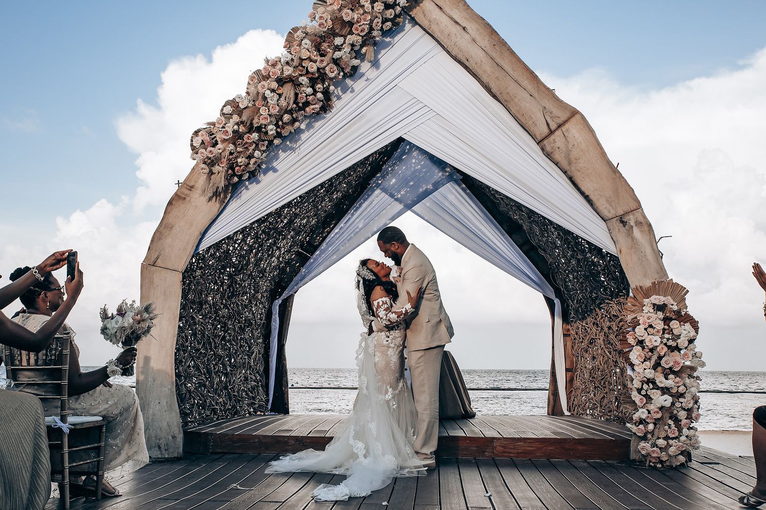 Wedding ceremony setup on a beachfront deck with white arch and black backdrop decorated with pink floral arrangements.