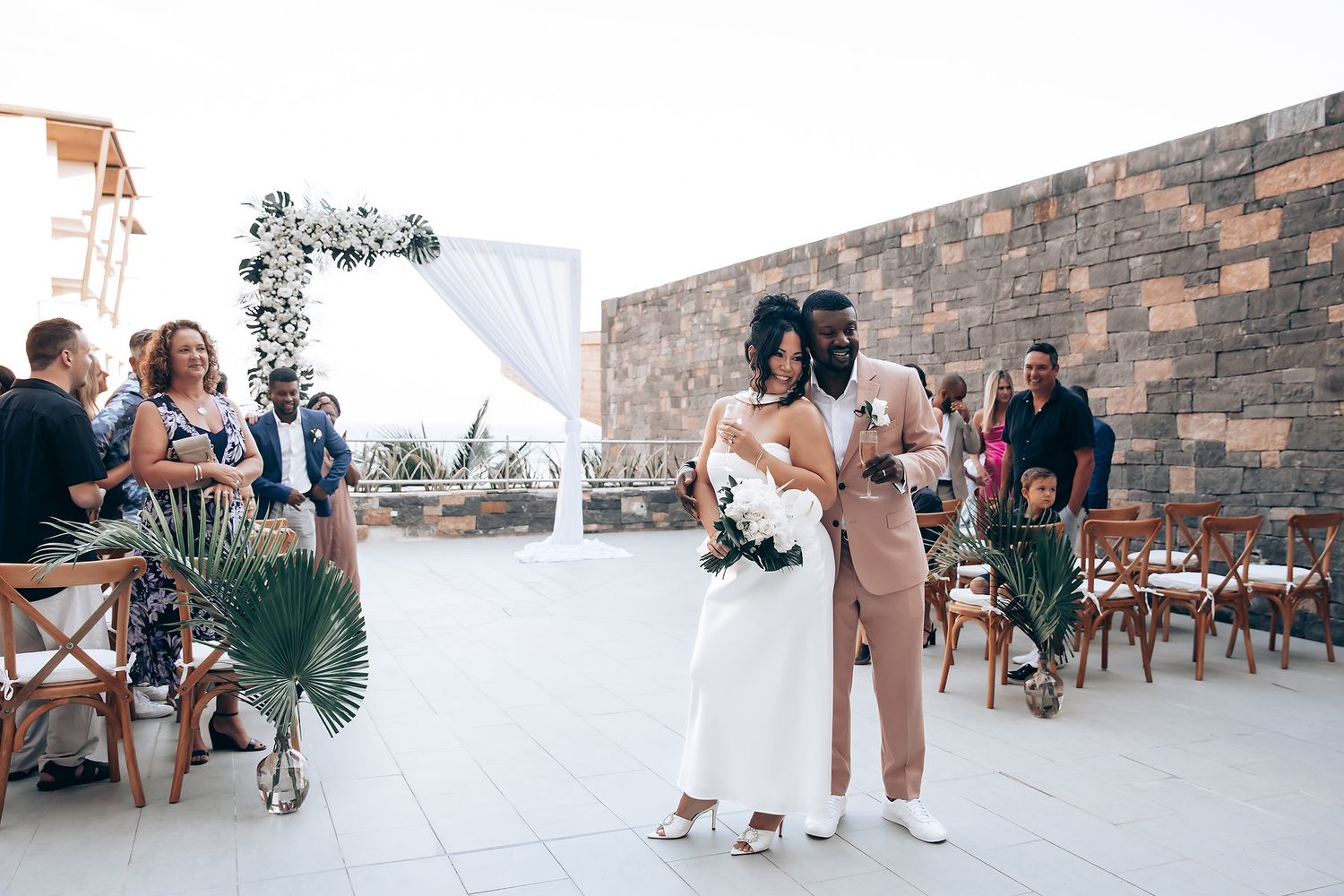 Wedding guests gather on an outdoor terrace with stone walls and white drapery.