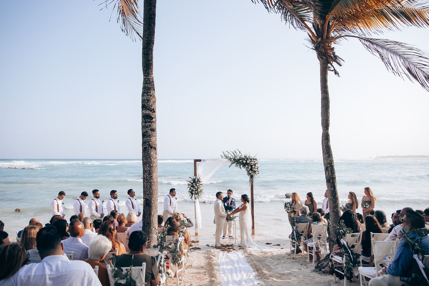 Wedding guests seated on white chairs at a tropical beach ceremony with palm trees and ocean views.