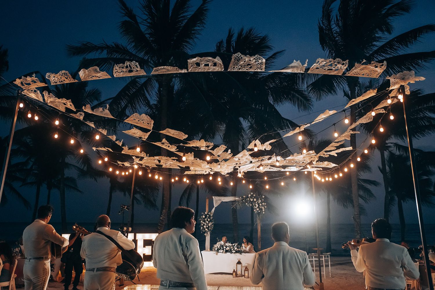 String lights illuminate an outdoor beach wedding reception at dusk with palm trees silhouetted against the dark sky.