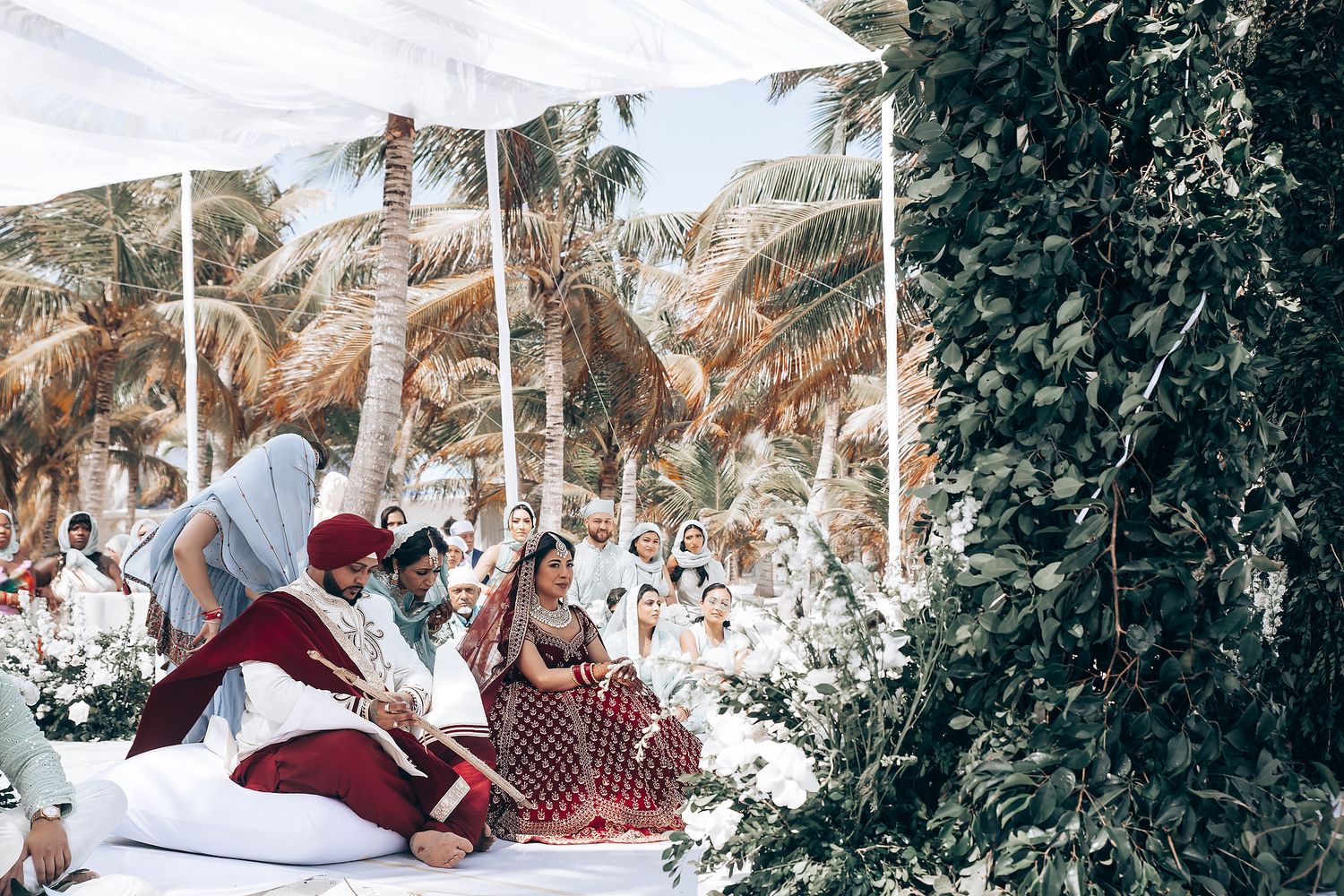Outdoor wedding celebration with guests seated under white canopy and palm trees.