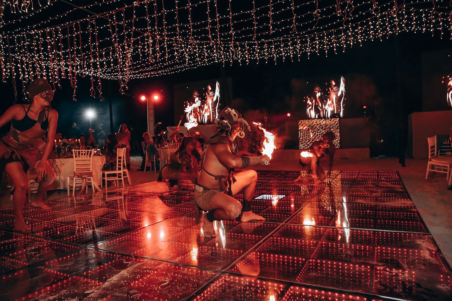 Fire dancers perform a mesmerizing nighttime show with flaming torches on a beach under string lights.