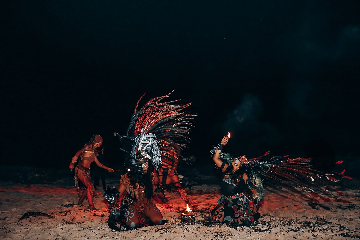 Silhouetted figures dance around a glowing red campfire against a dark night sky in a dramatic scene.