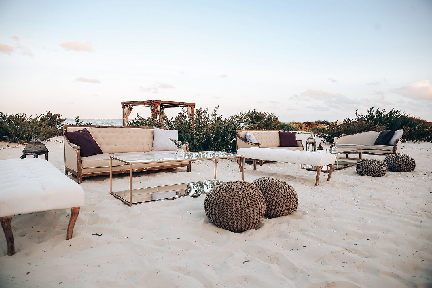 Beige wooden lounge chairs and woven poufs arranged on a sandy beach at sunset.
