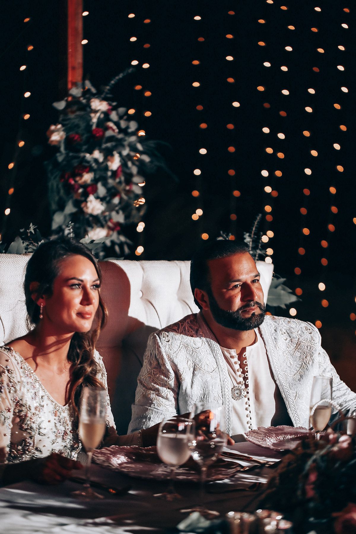 Four candid reception photos show guests in white attire seated at a table with twinkling lights in the background.