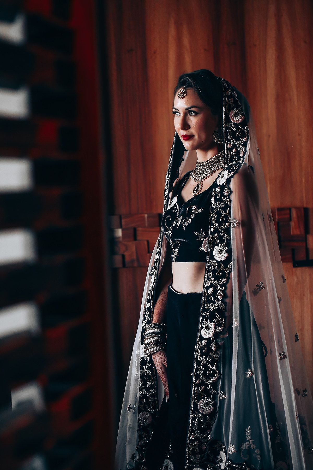 A dramatic bridal portrait in black attire with a flowing veil against a dark wooden backdrop.