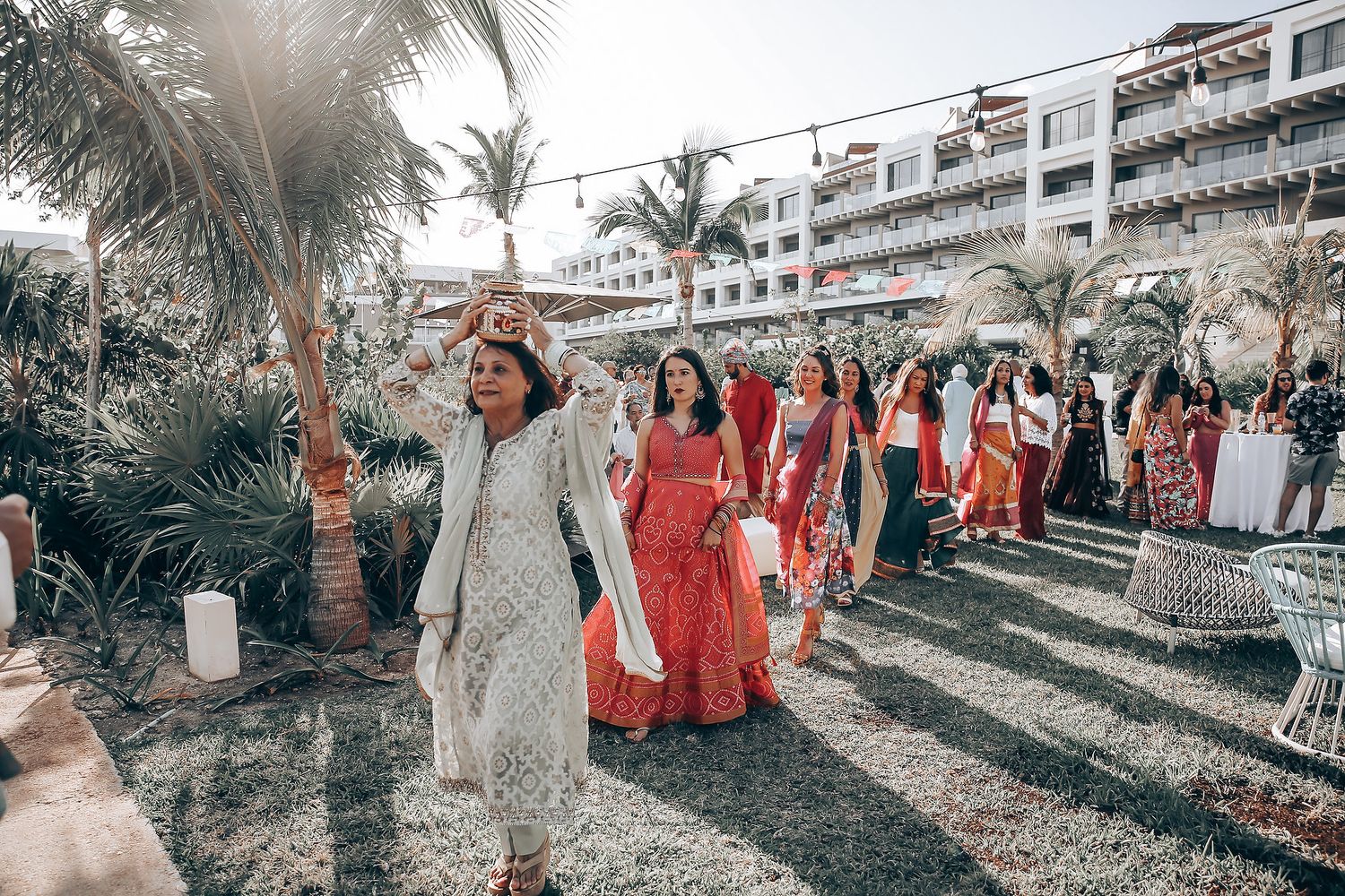 Wedding party in formal attire walks along palm tree-lined path during outdoor celebration.