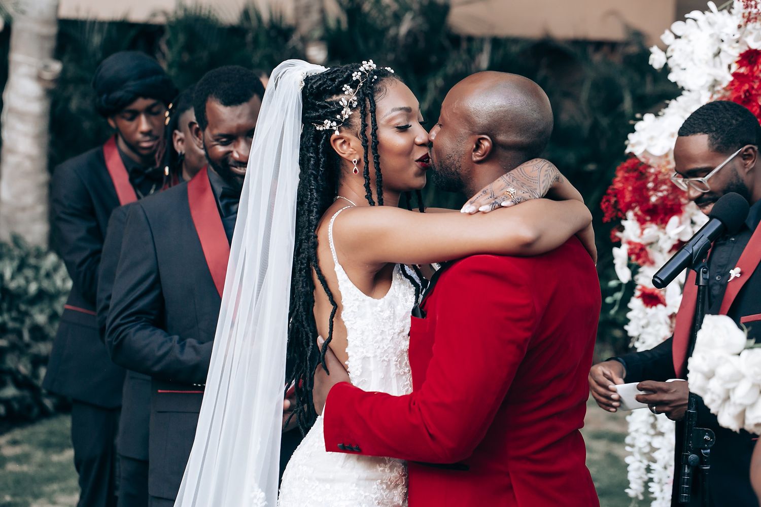 A bride and groom share a romantic kiss during their wedding ceremony with red and white floral decor in the background.
