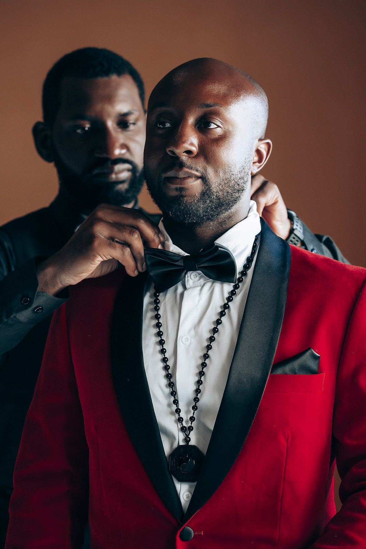 Two people in formal attire share an intimate moment wearing red blazers and black necklaces against a dark background.