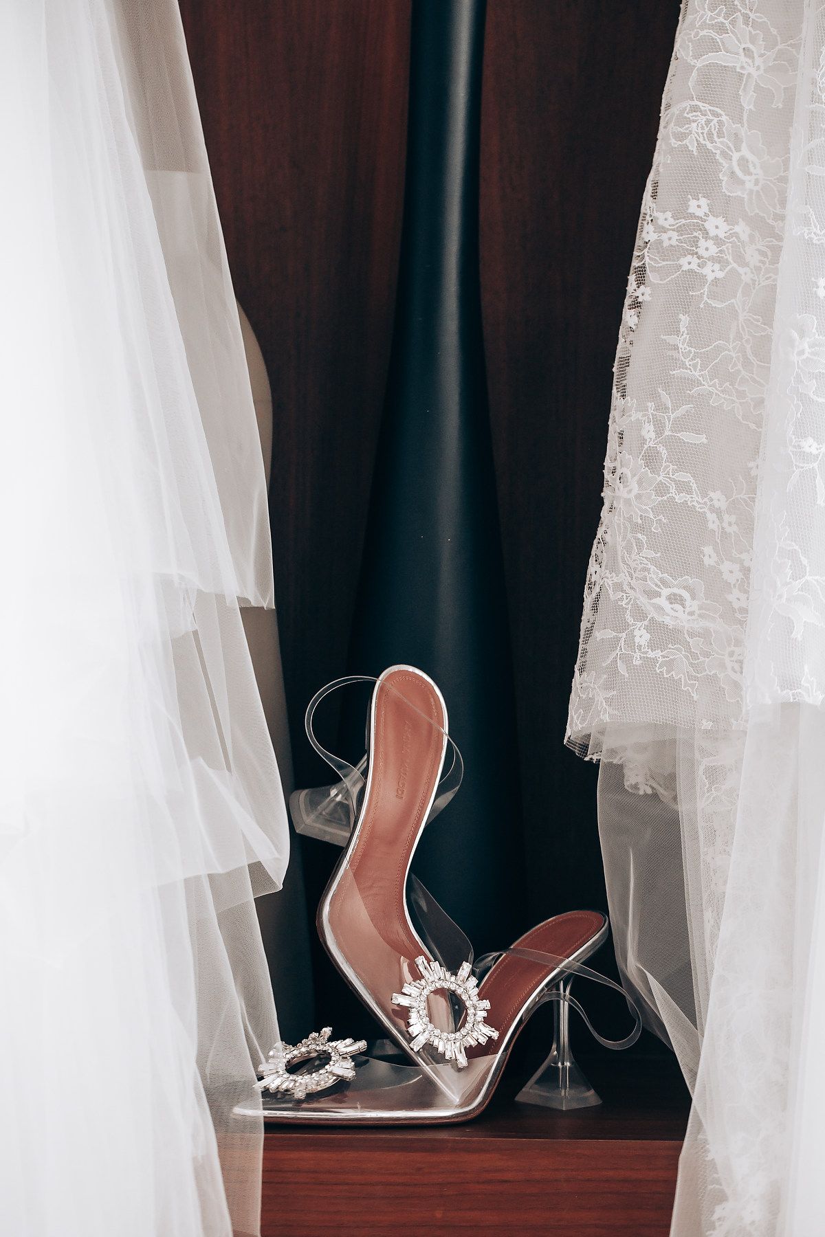 Elegant bridal shoes with crystal embellishments sit between white lace curtains on a dark wooden surface.