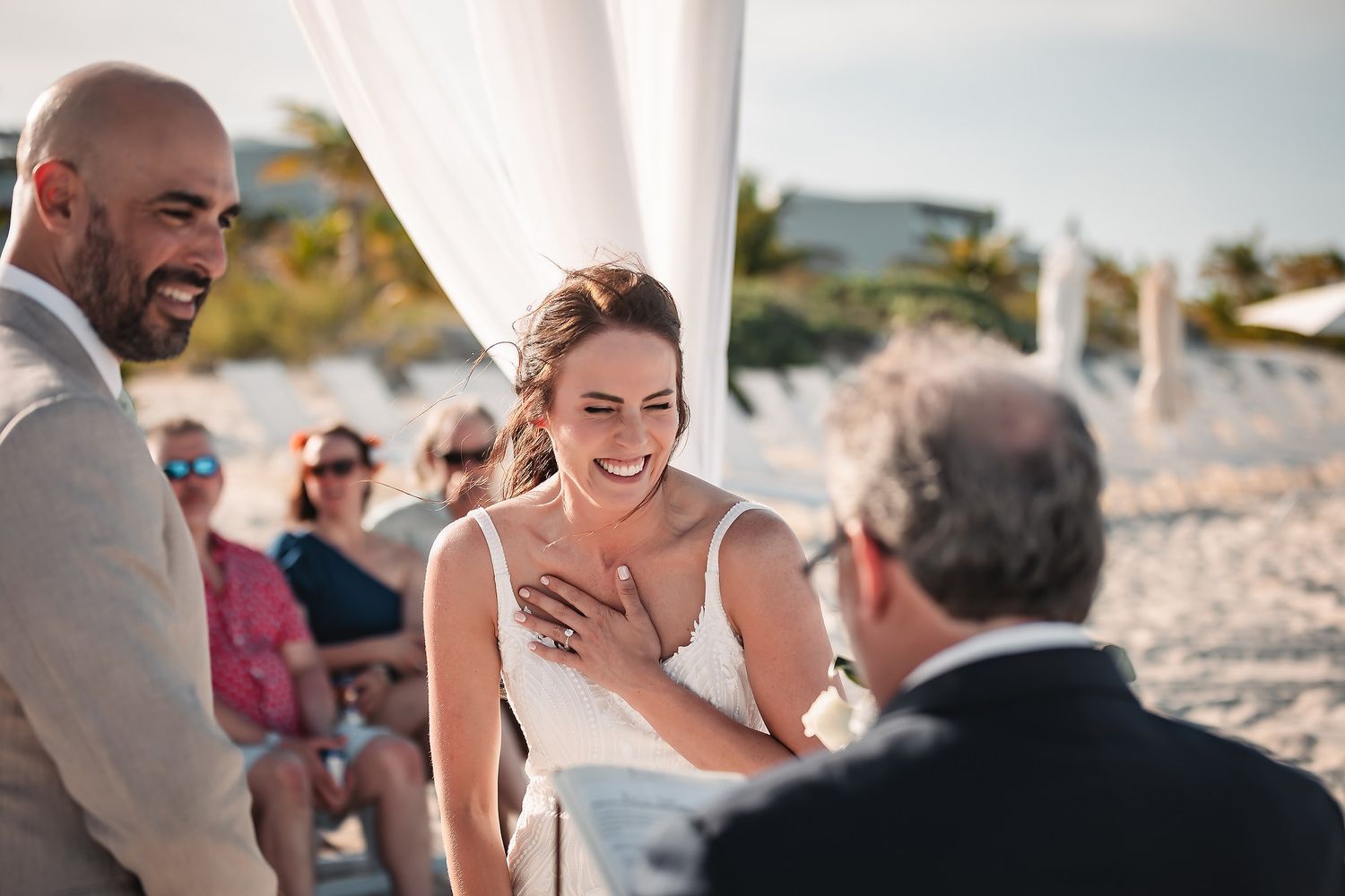 A beach wedding ceremony takes place with guests seated on the sand during golden hour.
