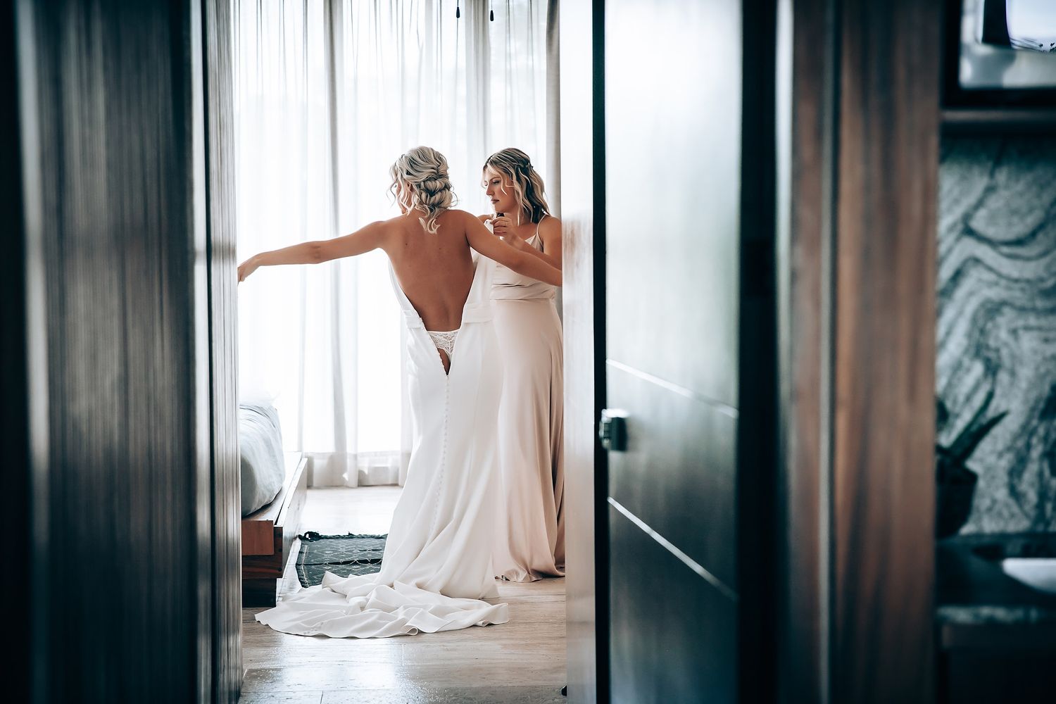 Wedding dress fitting scene captured through doorway with natural lighting.