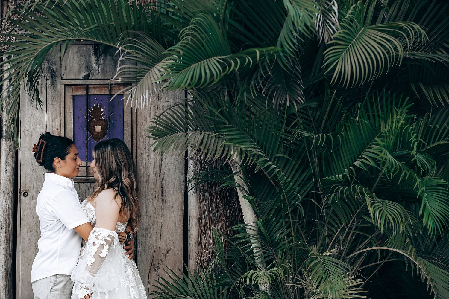 A couple shares an intimate moment against a wooden door surrounded by tropical foliage.