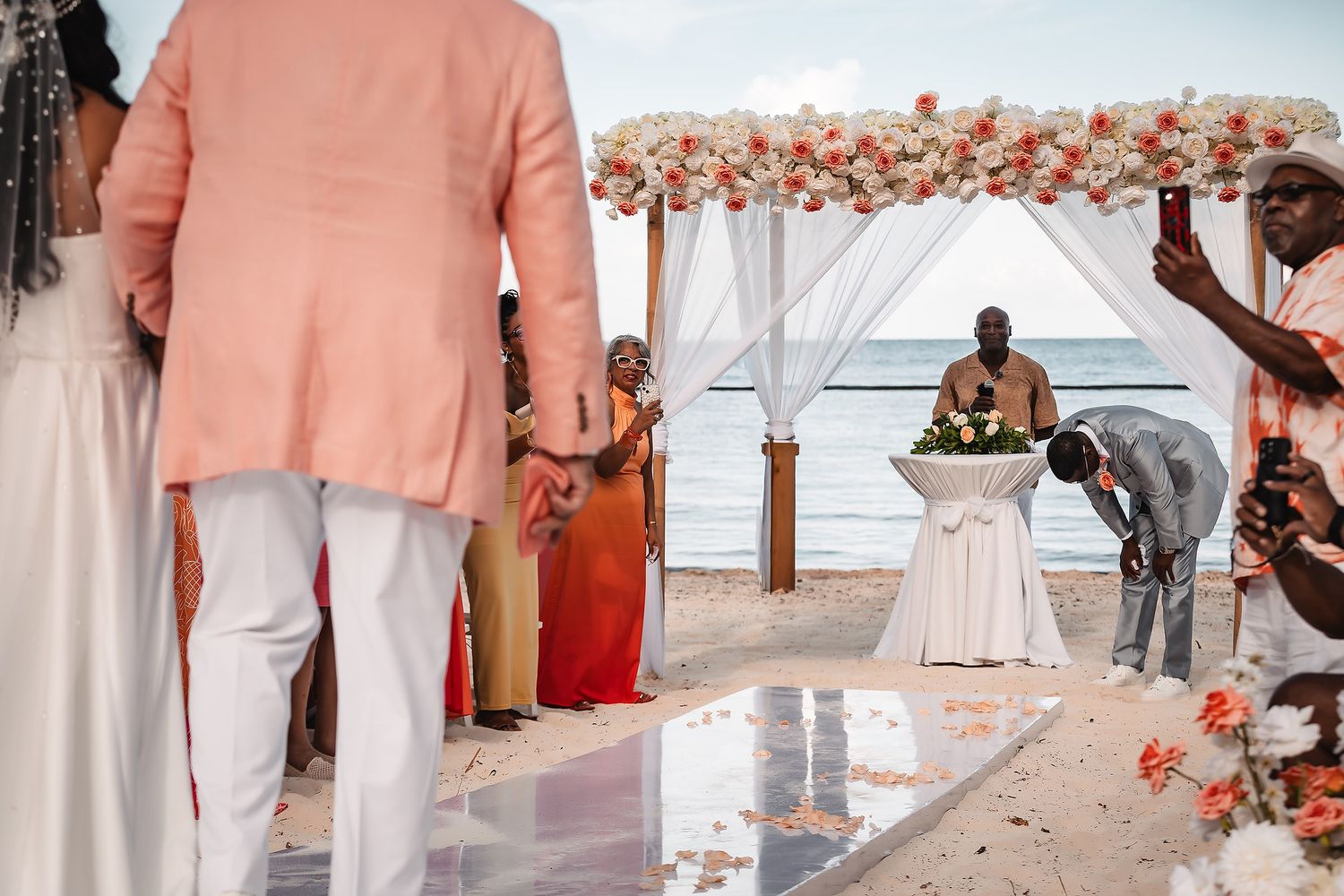 Beach wedding ceremony setup with white canopy and coral floral decorations against an ocean backdrop.