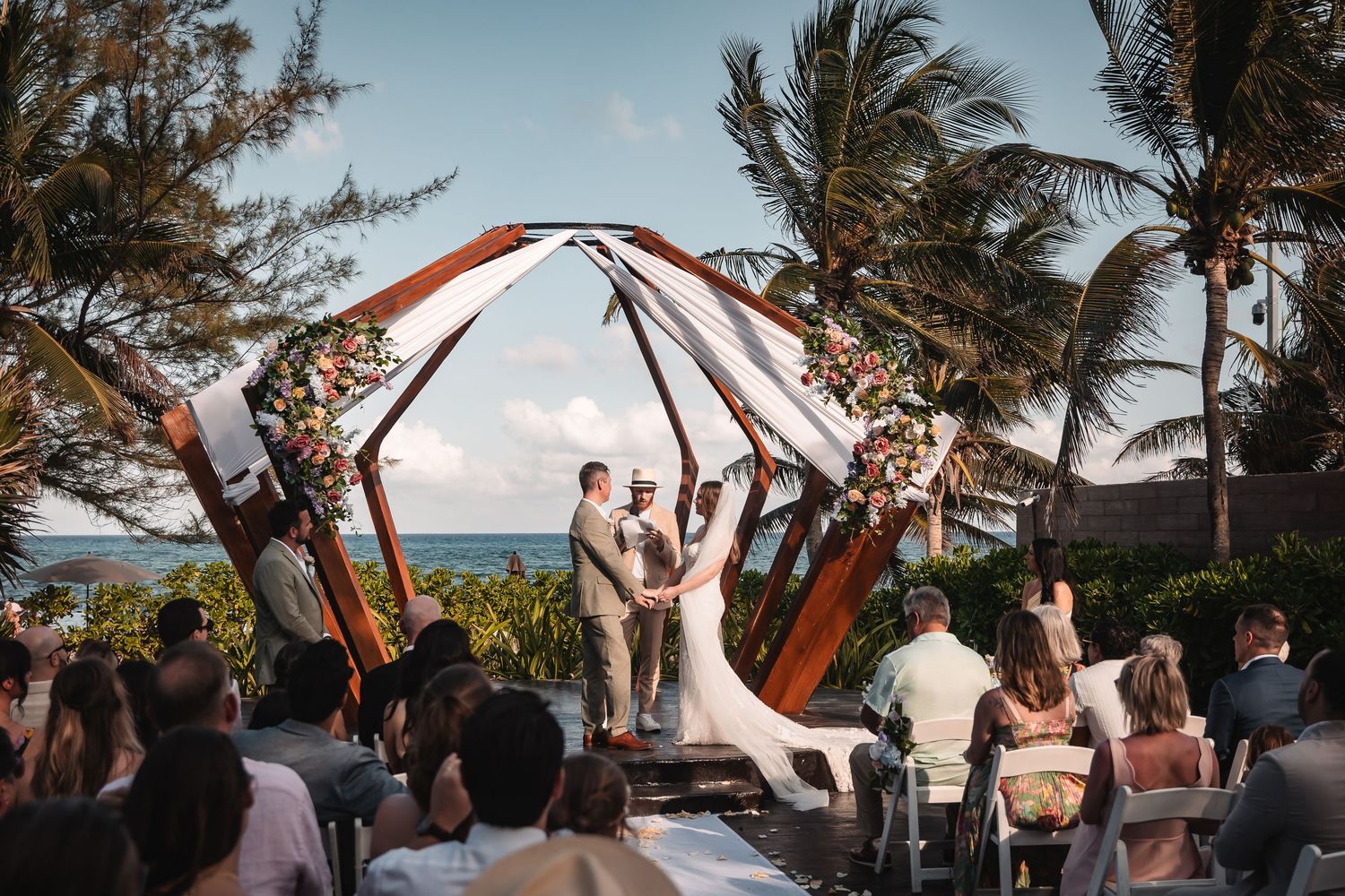 Beachfront wedding ceremony with guests gathered around wooden pergola structure.