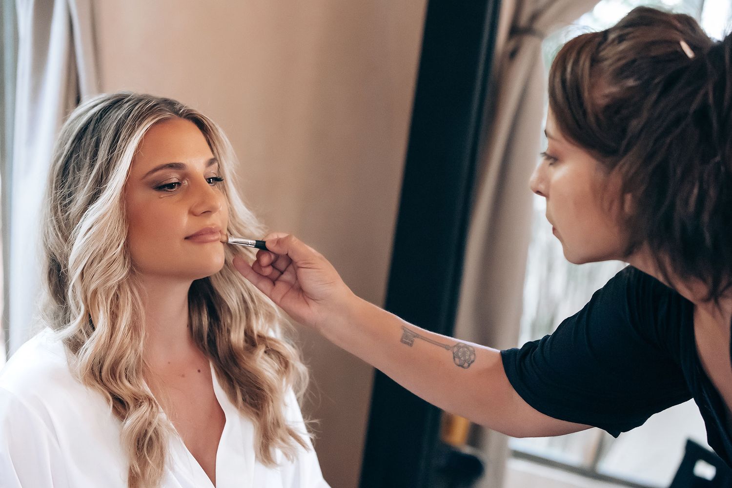 Wedding preparation scene with makeup being applied in natural window light.