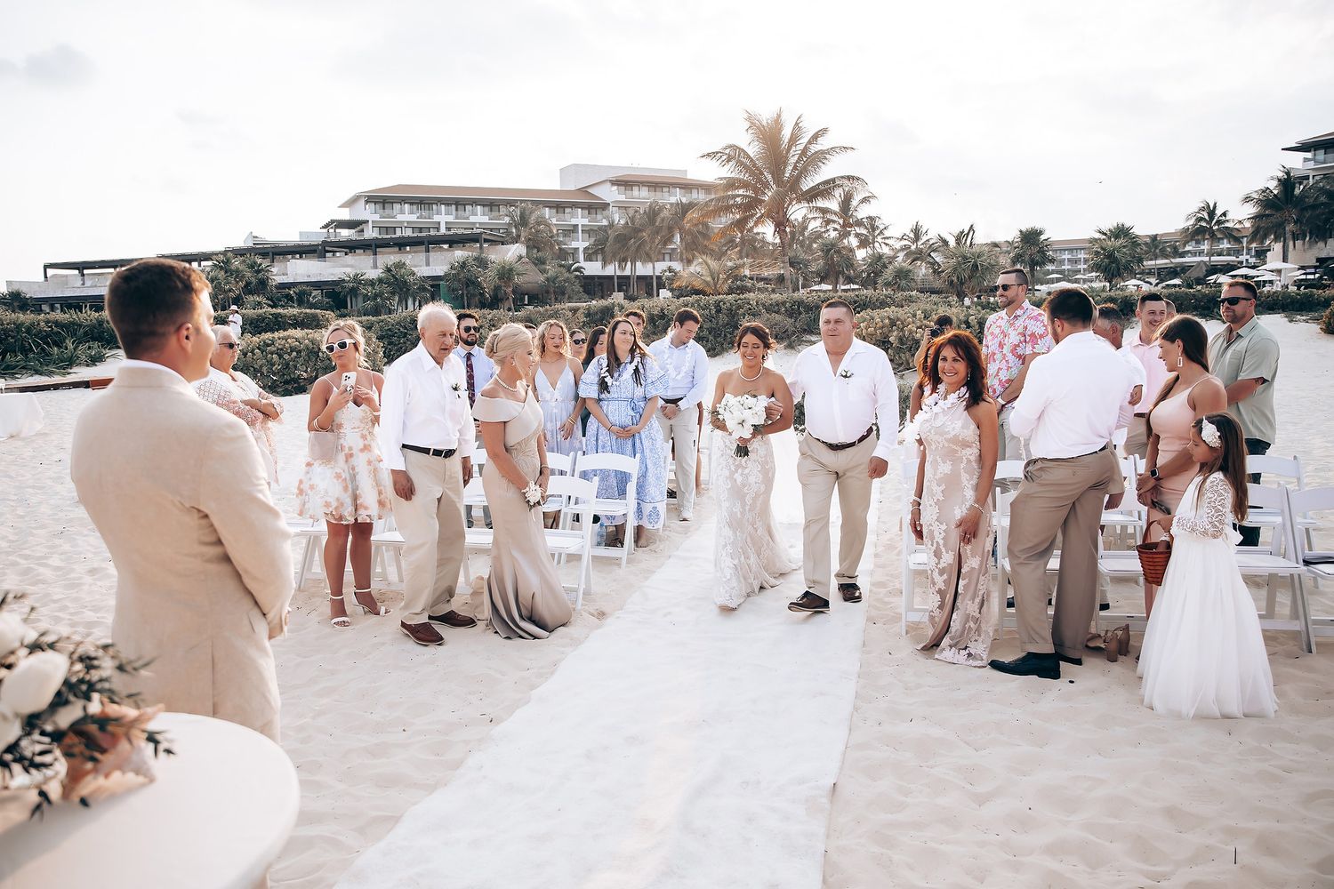 Wedding guests in white and beige attire gather on a tropical beach for a destination ceremony.