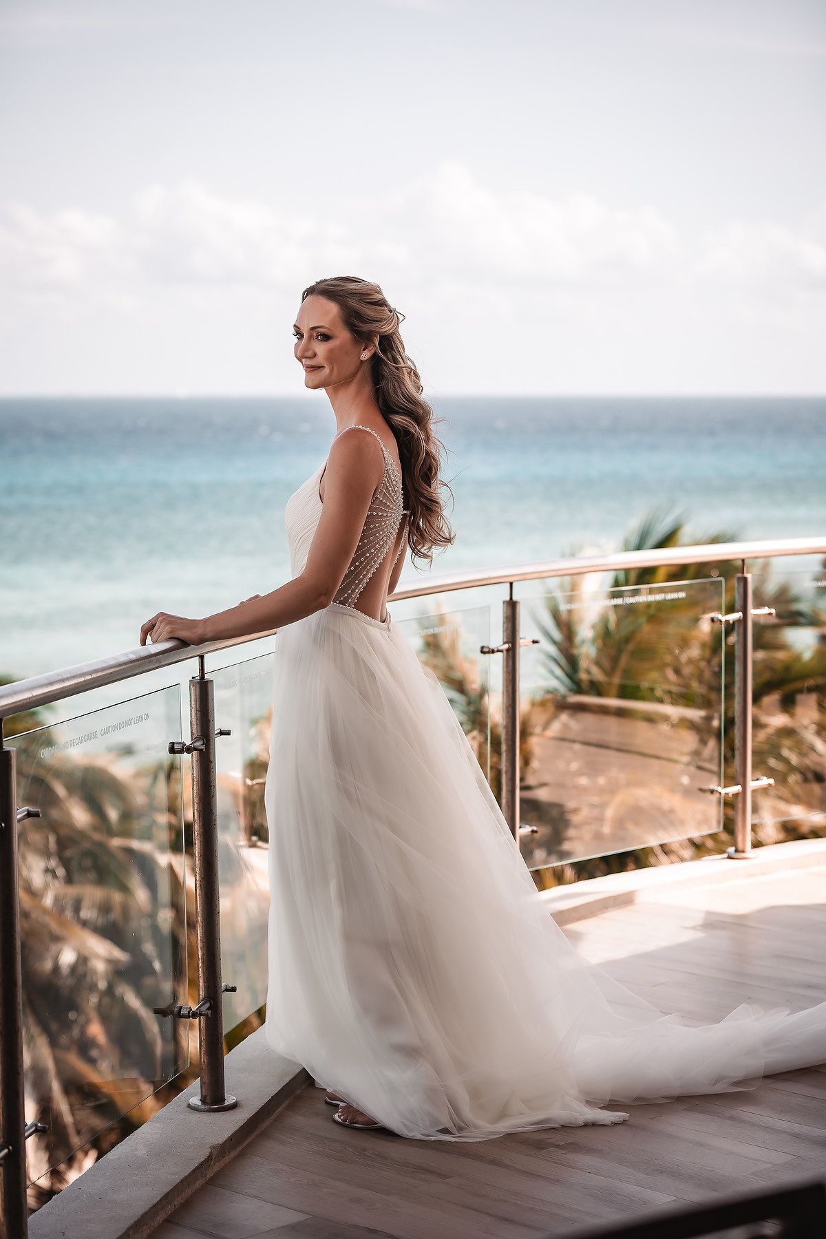 Bride in flowing white gown standing on oceanfront balcony with palm trees in background.
