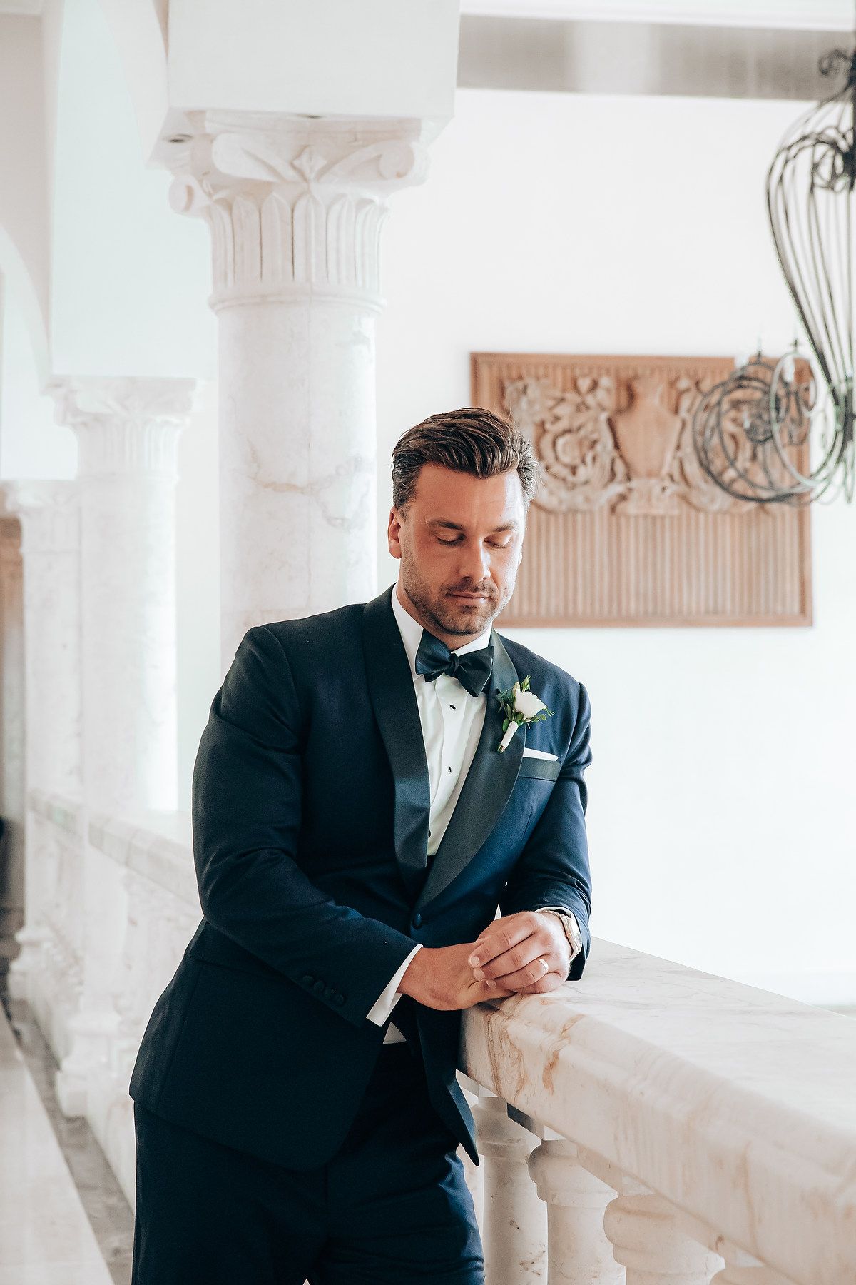 A groom in a dark suit leans thoughtfully against an elegant white column in a bright, classical interior.