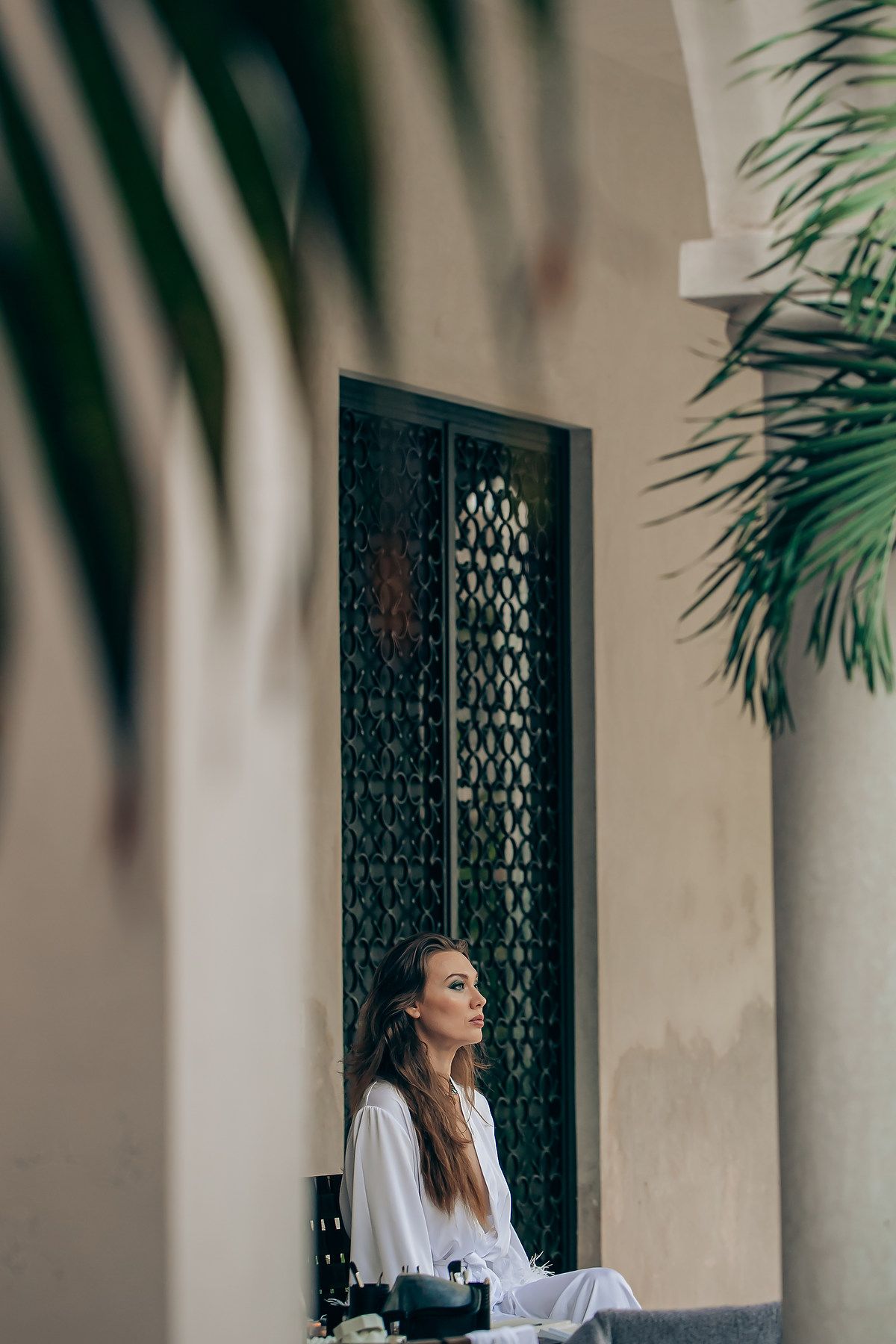 Person in white sitting by ornate window with palm fronds creating dramatic shadows in elegant interior setting.