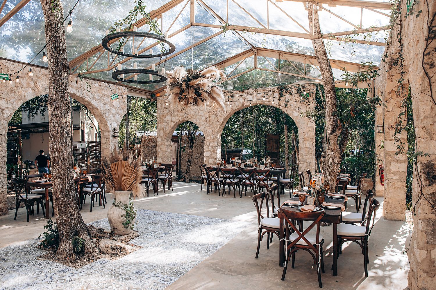 Rustic outdoor dining area with stone arches, wooden tables, and natural trees growing through a glass-covered patio.