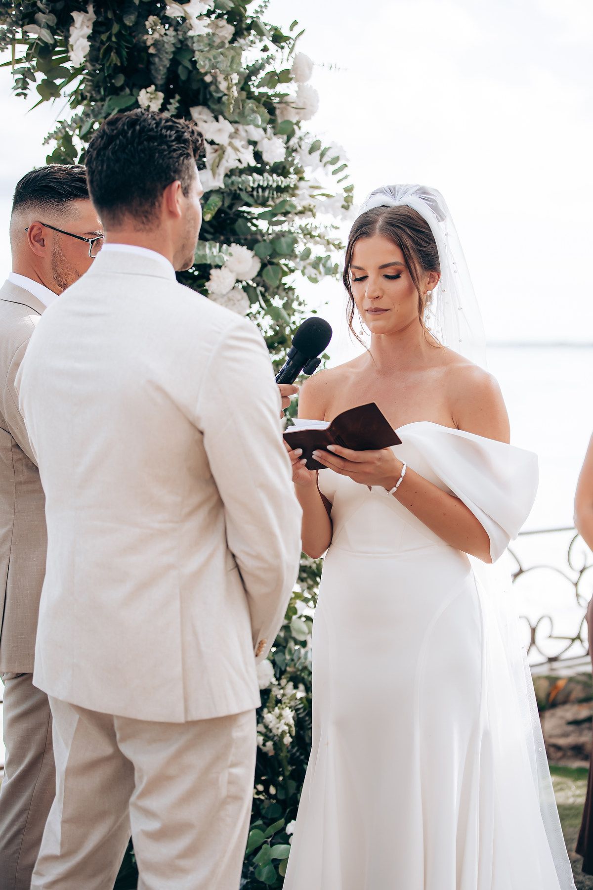 Wedding ceremony attendees in white attire gathered outdoors under floral decorations on a sunny day.