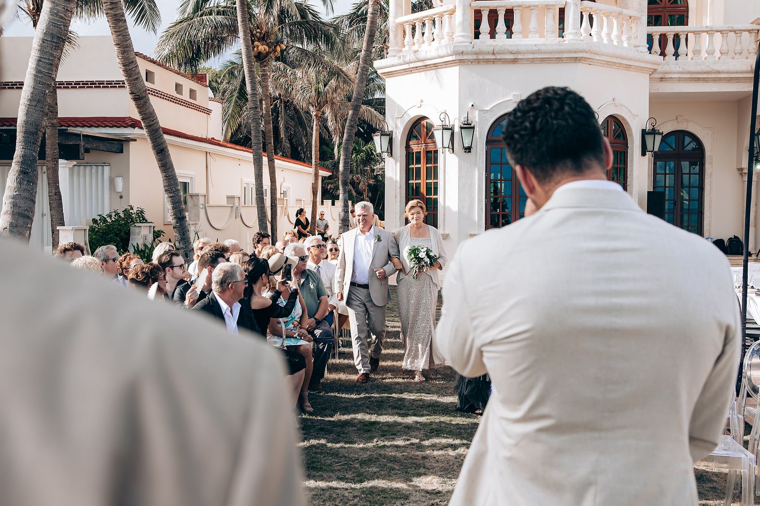 A wedding ceremony takes place outdoors at an elegant venue with palm trees and white Mediterranean-style architecture.