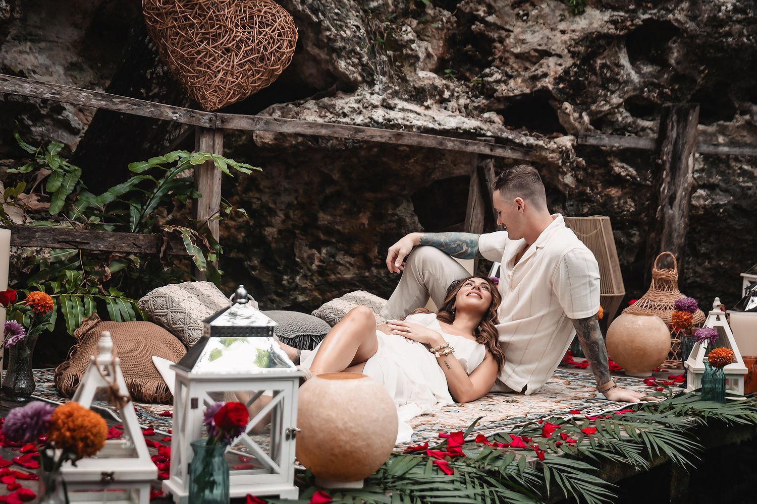 Romantic outdoor setup with lanterns, pillows and rose petals scattered on a decorative rug in a natural setting.