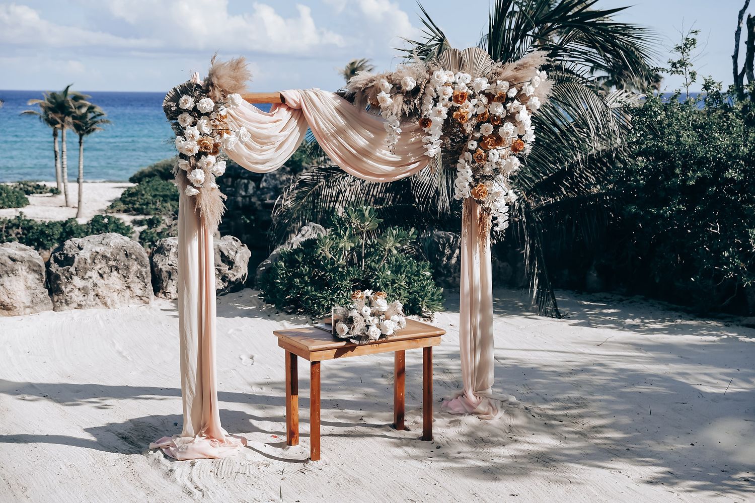 Beachfront wedding ceremony setup with blush floral arch and white chairs overlooking the ocean.