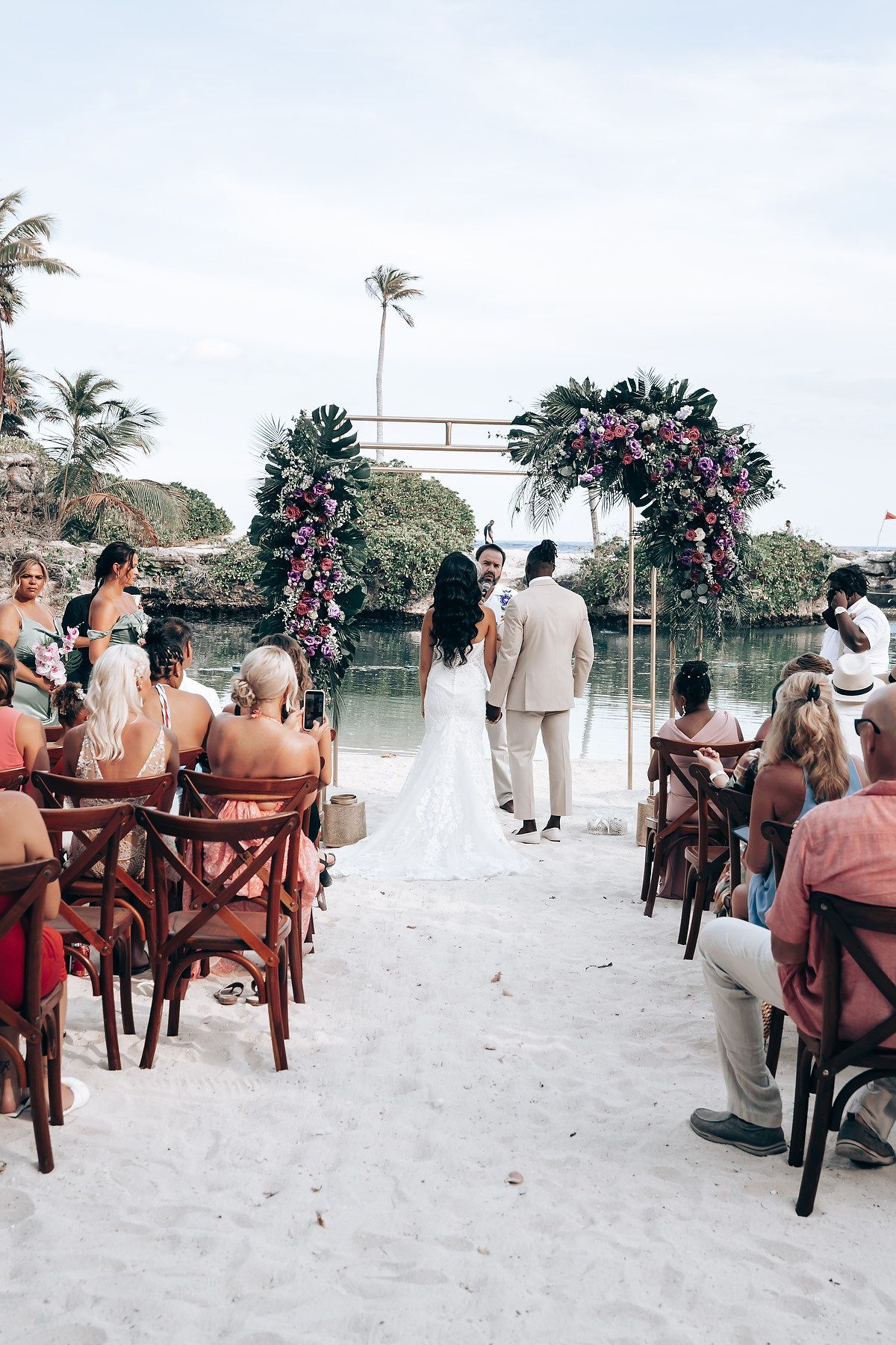 Beach wedding ceremony setup with wooden chairs and floral arrangements on white sand.