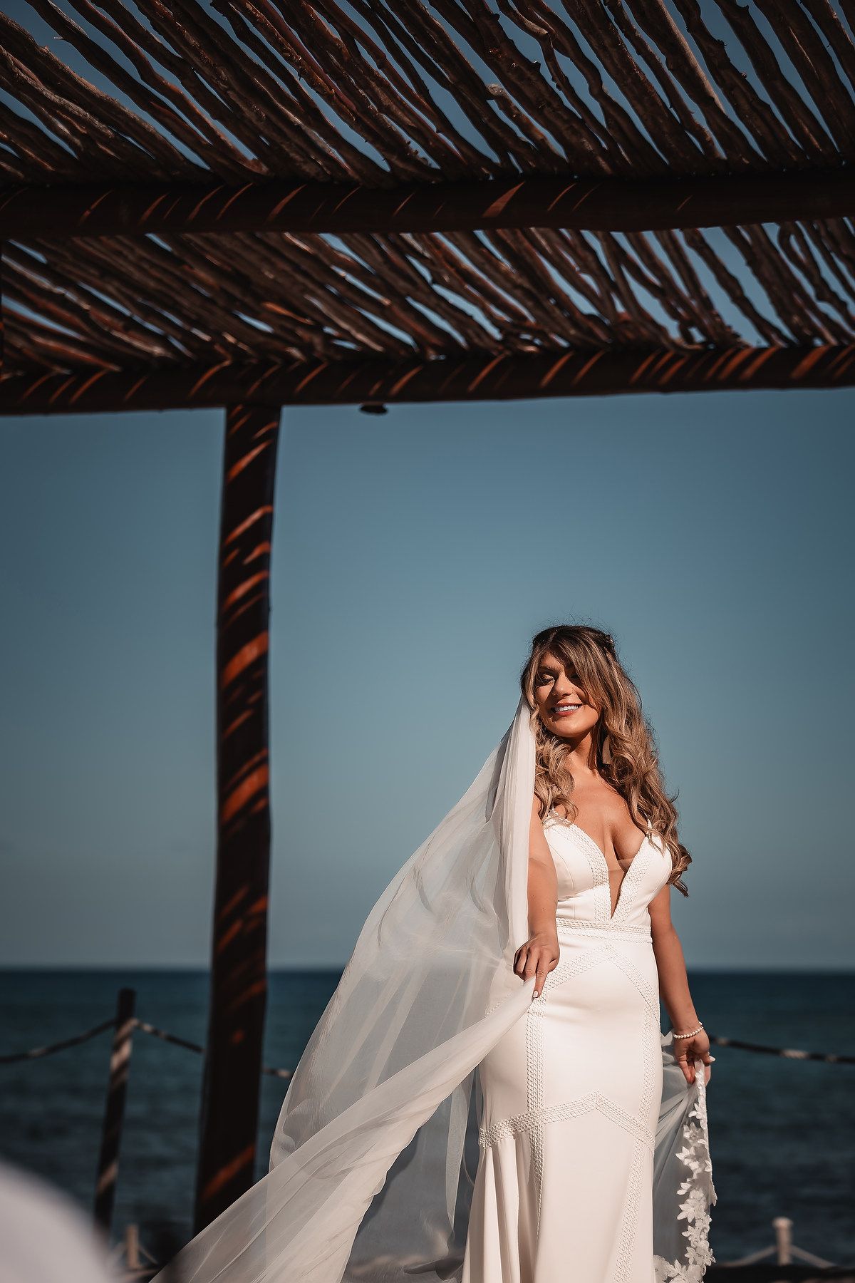 A flowing white wedding dress and veil against an ocean backdrop under a thatched roof.