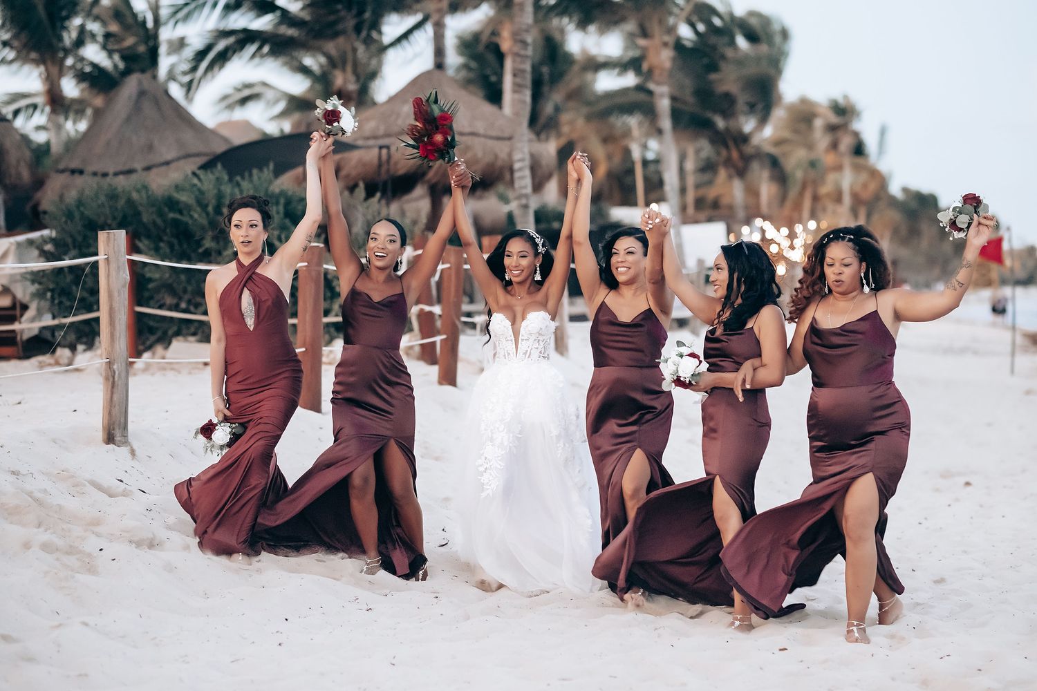 Wedding party members in burgundy and white attire celebrate on sandy beach.