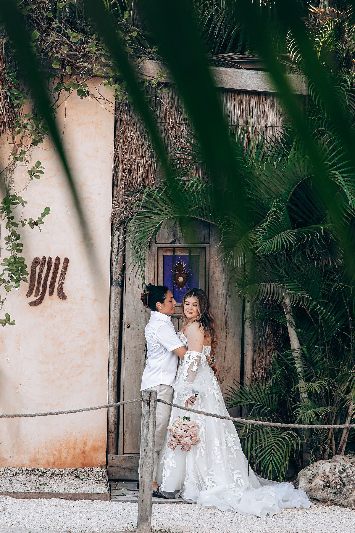 A romantic wedding photoshoot in a tropical garden setting with white walls and palm fronds.